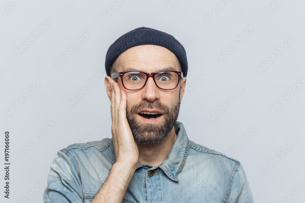 Indoor shot of surprised handsome man stares at camera, keeps hand on cheek, wears glasses and stylish clothing, isolated over white studio backgrpund. People, facial expressions and emotions Indoor shot of surprised handsome man stares at camera, keeps hand on cheek, wears glasses and stylish clothing, isolated over white studio backgrpund. People, facial expressions and emotions