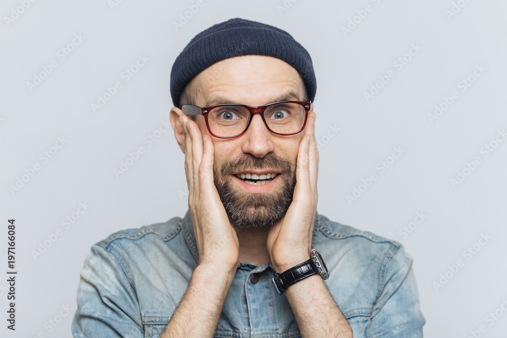 Indoor shot of excited bearded man with blue eyes and cheerful expression, keeps hands on cheeks, dressed in fashionable clothing, isolated over white background. People, facial expressions concept Indoor shot of excited bearded man with blue eyes and cheerful expression, keeps hands on cheeks, dressed in fashionable clothing, isolated over white background. People, facial expressions concept