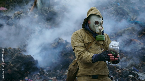 Fototapeta Naklejka Na Ścianę i Meble -  a man in a gas mask holds a sprout in the flask, against the background of fuming debris
