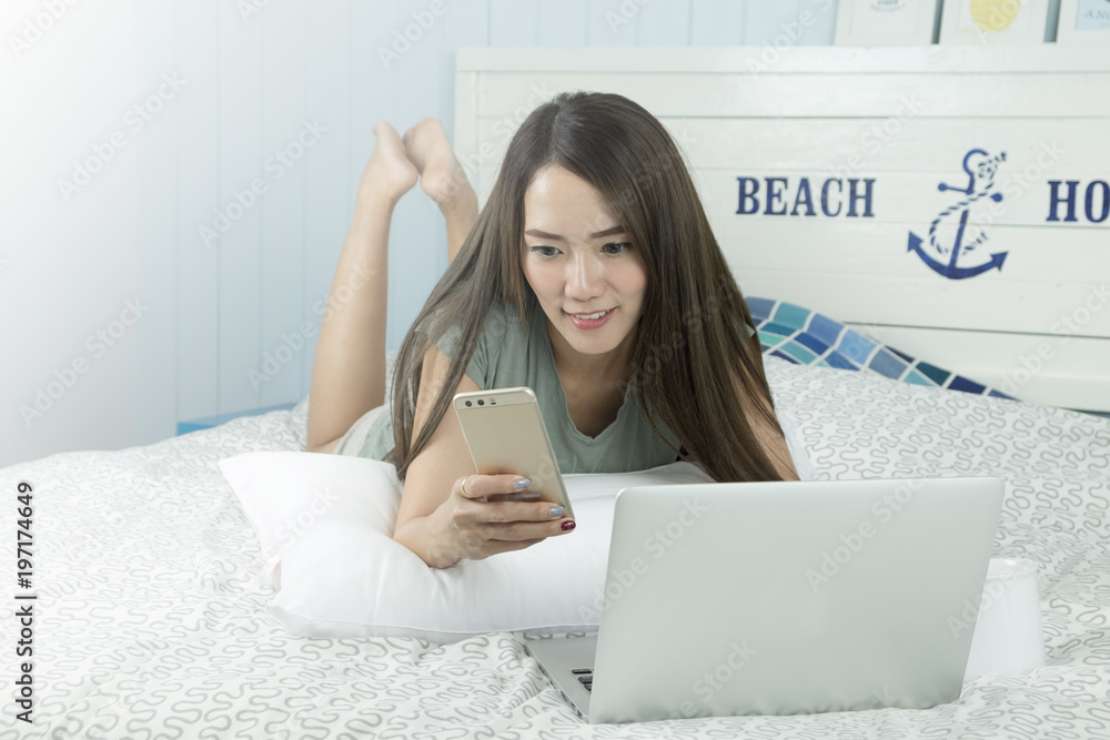Attractive smiling asian woman using laptop and smartphone while lying on her bed. Attractive smiling asian woman using laptop and smartphone while lying on her bed.