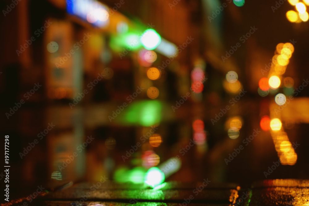 Night city lights bokeh from the shopping center, shop windows. Colorful surface of asphalt, blurred background. 