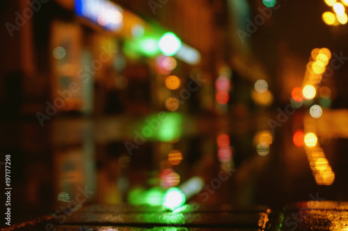 Night city lights bokeh from the shopping center, shop windows. Colorful surface of asphalt, blurred background. 