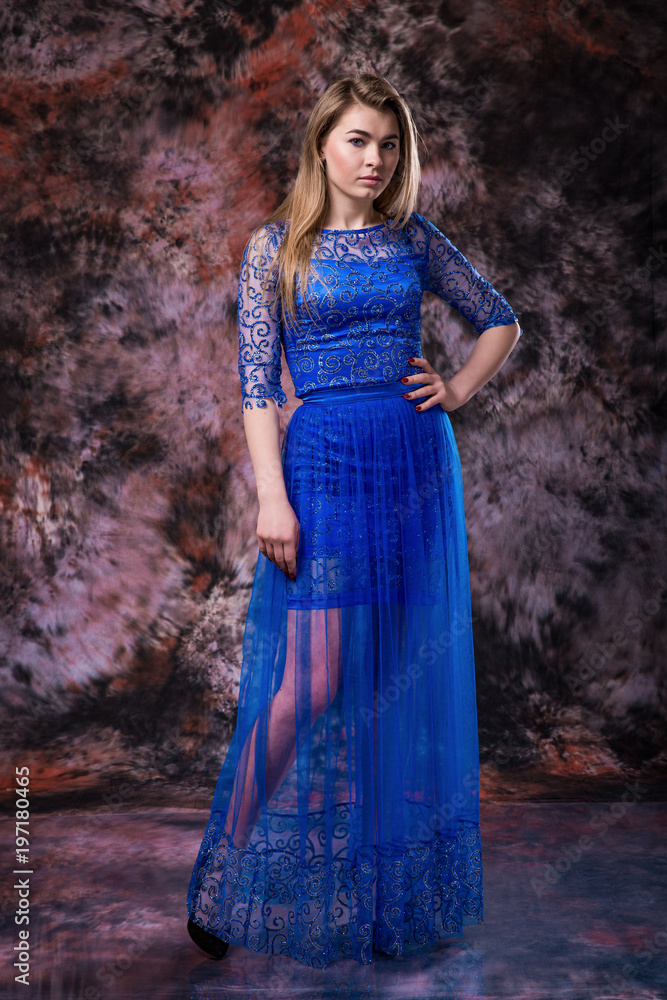 Elegant blonde woman in the blue evening sparkling dress is posing on marble colored background Elegant blonde woman in the blue evening sparkling dress is posing on marble colored background