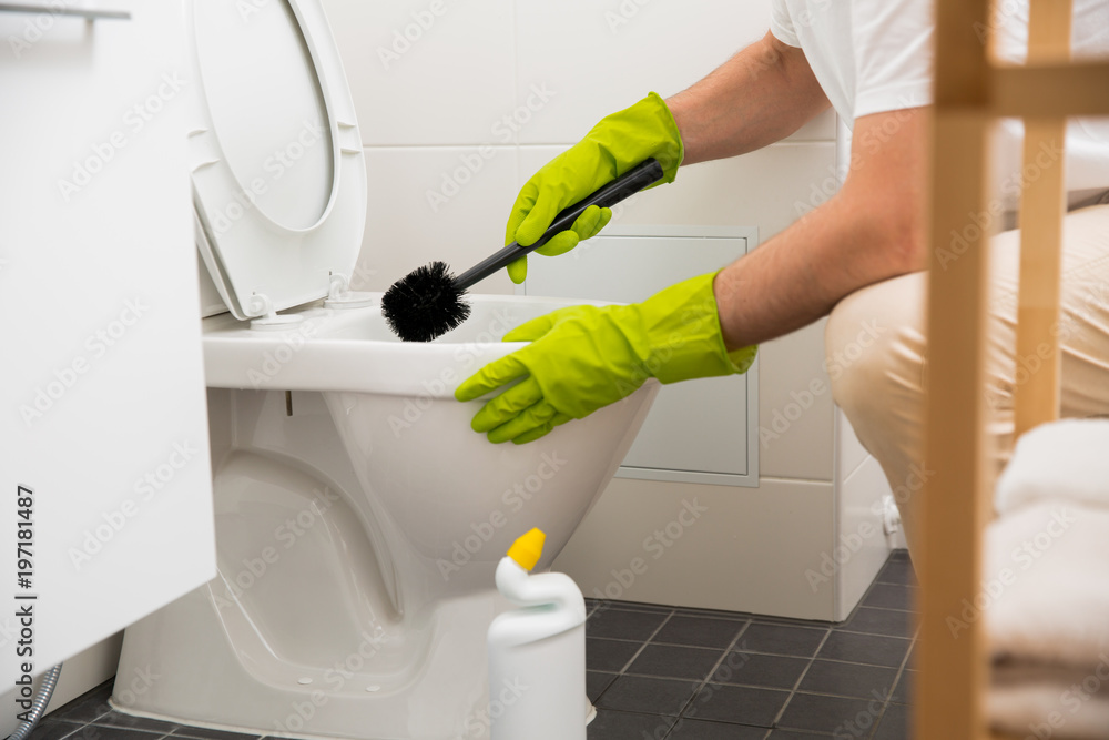 Man in rubber protective gloves cleaning and washing toilet with brush and bottle of cleaning agent in hands. White modern bathroom. Green liquid detergent dripping in toilet pan Man in rubber protective gloves cleaning and washing toilet with brush and bottle of cleaning agent in hands. White modern bathroom. Green liquid detergent dripping in toilet pan