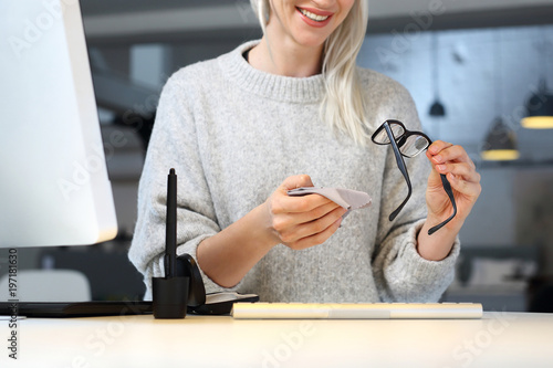 Okulary do pracy przy komputerze. Kobieta siedzi przy biurku i wyciera okulary ściereczką.