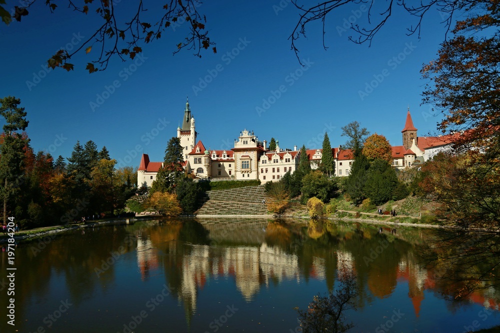 Fototapeta premium Beautiful famous Pruhonice castle in Czech Republic, Europe standing on hill, view over pond with reflection in water, spring landscape, trees, plants, clear dark blue sky, people walking