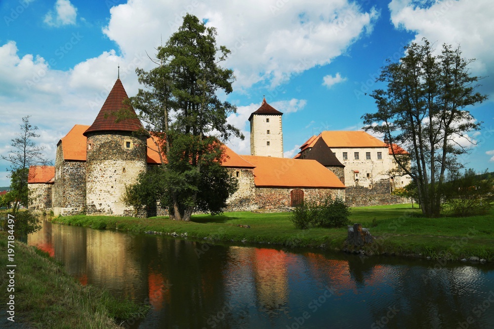 Famous water castle Svihov at Czech republic, summer picture with moat, reflection in water, trees, green grass, blue sky, white clouds, palace, church, red roofs, rampart, beautiful scenery