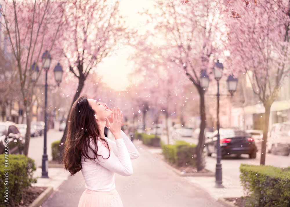 Young woman enjoying cherry tree petals falling to her face, happy emotion pink pastel colors, sunflare in background Young woman enjoying cherry tree petals falling to her face, happy emotion pink pastel colors, sunflare in background