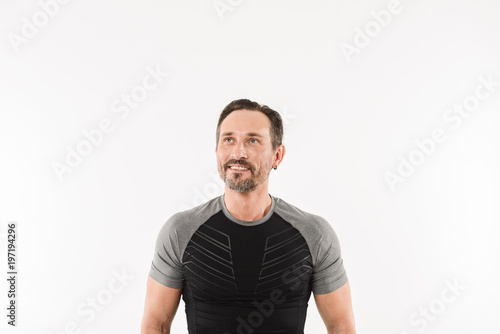 Portrait of masculine man 30s wearing sportswear looking upward on copyspace with smile and satisfied look after doing sports, isolated over white background