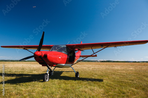 A small red light-engine aircraft with a propeller stands in the middle of a field with grass on a summer clear sunny day.