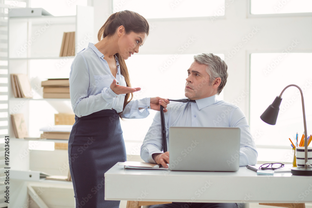 Fototapeta premium Relations between colleagues. Nice pleasant young woman holding a mans tie and talking to him while standing in the office