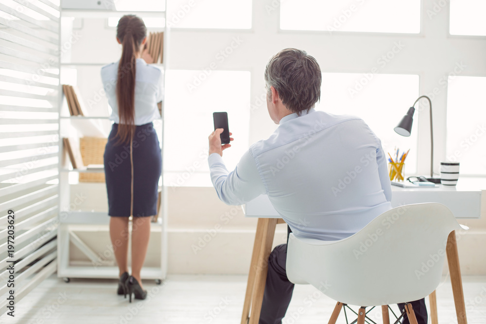 Inappropriate photos. Unpleasant nice adult man holding his smartphone and looking at his female colleague while taking photos Inappropriate photos. Unpleasant nice adult man holding his smartphone and looking at his female colleague while taking photos