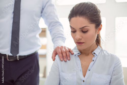 Female abuse. Nice cheerless unhappy woman sitting in the office and looking at the male hand while being sexually harassed photo