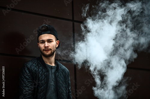 Vape man. Portrait of a handsome young white guy in a modern black cap vaping and letting off puffs of steam from an electronic cigarette opposite the futuristic urban background. © aleksandr_yu