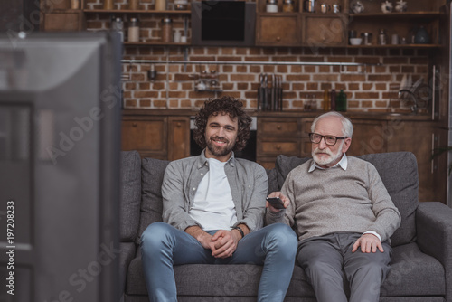 adult son and senior father watching tv on sofa at home