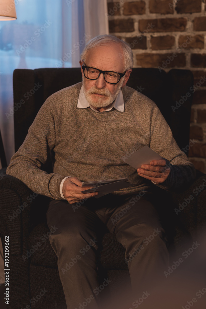 senior  man sitting in armchair, holding old photos and looking away