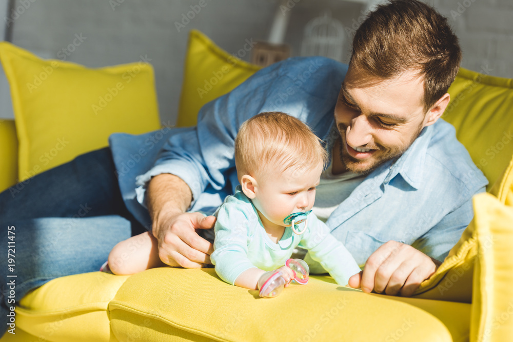 Smiling father and infant daughter with baby dummy on sofa Smiling father and infant daughter with baby dummy on sofa