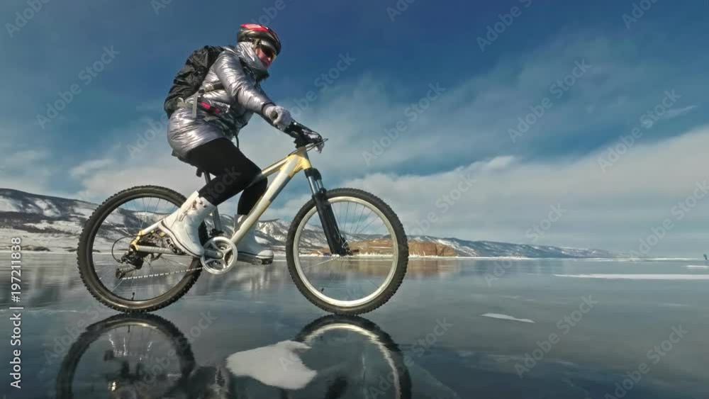 Woman is riding bicycle on the ice. The girl is dressed in a silvery down jacket, cycling backpack and helmet. Ice of the frozen Lake Baikal. The tires on the bicycle are covered with special spikes Woman is riding bicycle on the ice. The girl is dressed in a silvery down jacket, cycling backpack and helmet. Ice of the frozen Lake Baikal. The tires on the bicycle are covered with special spikes