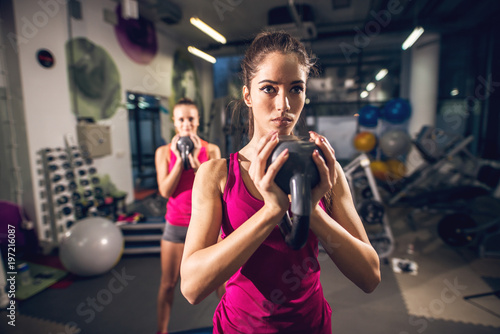 Two young shape athletic cute attractive slim fitness active girls doing exercises with kettlebells while holding upside down in the modern gym.