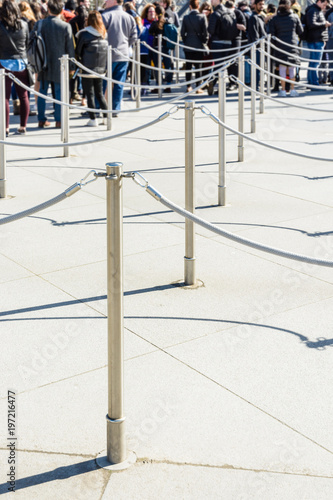 Stainless steel poles linked by grey ropes for queue control at the entrance of a tourist site, with drop shadows on a light ground and blurry people queueing up in the background.