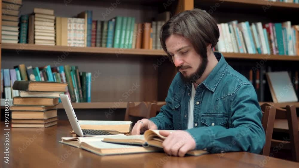 Hard-working caucasian student studying in library, young man with beard and straight fair hair using laptop while reading book, sitting at brown table next to the bookshelves Hard-working caucasian student studying in library, young man with beard and straight fair hair using laptop while reading book, sitting at brown table next to the bookshelves
