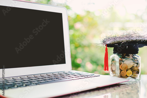 Computer laptop and square academic cap with the glass jar of coin against blurred natural green background