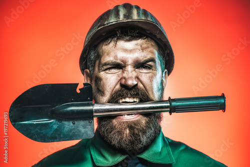 Mining and construction concept - man with angry face in protective helmet holds shovel in teeth. Bearded man wears working uniform and harthat. Man worker in helmet holds sapper shovel Military spade photo