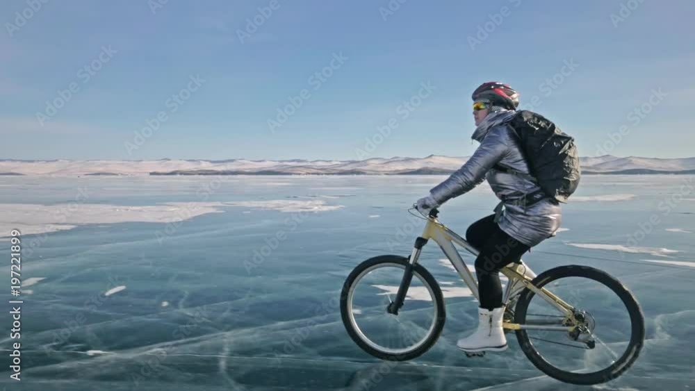 Woman is riding bicycle on the ice. The girl is dressed in a silvery down jacket, cycling backpack and helmet. Ice of the frozen Lake Baikal. The tires on the bicycle are covered with special spikes Woman is riding bicycle on the ice. The girl is dressed in a silvery down jacket, cycling backpack and helmet. Ice of the frozen Lake Baikal. The tires on the bicycle are covered with special spikes