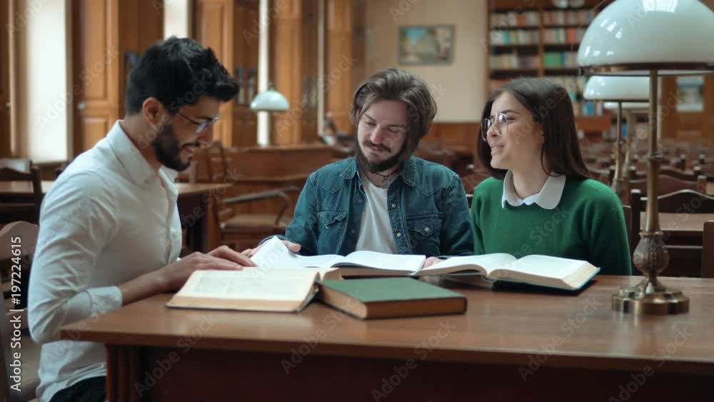 Three intelligent students are sitting in big library, chatting about subject while looking through books, beautiful woman wearing green sweater, man near by - denim shirt, black-haired man - white Three intelligent students are sitting in big library, chatting about subject while looking through books, beautiful woman wearing green sweater, man near by - denim shirt, black-haired man - white