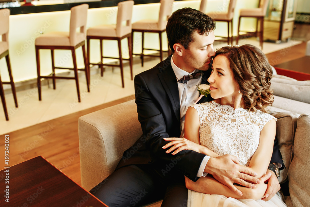Bride and groom sitting at the bar on the couch. Cute married couple in cafe, groom kissing a bride. Pure tenderness Bride and groom sitting at the bar on the couch. Cute married couple in cafe, groom kissing a bride. Pure tenderness