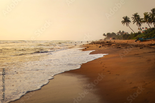 Empty beach during afternoon sun light, sand wet from waves, with silhouettes of people playing in background. Kalutara, Sri Lanka