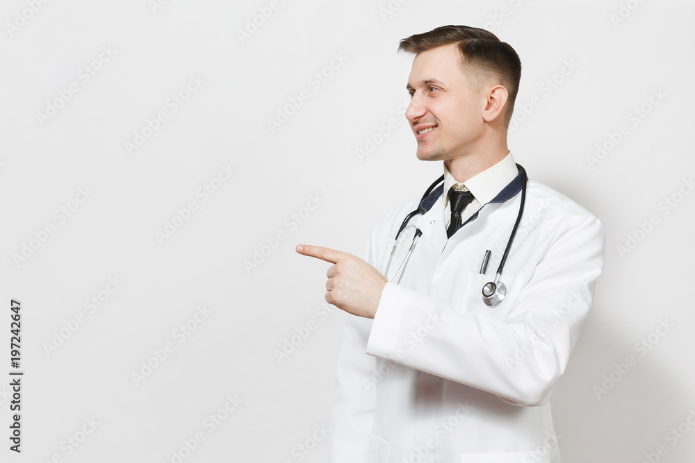 Smiling handsome young doctor man isolated on white background. Male doctor in medical gown, stethoscope pointing index finger aside on copy space. Healthcare personnel, health, medicine concept. Smiling handsome young doctor man isolated on white background. Male doctor in medical gown, stethoscope pointing index finger aside on copy space. Healthcare personnel, health, medicine concept.