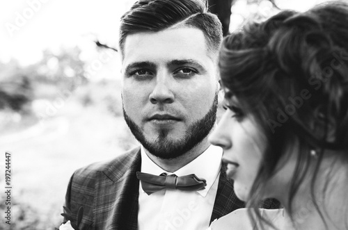 Beautiful wedding couple portrait in the forest. The bride with elegant hairstyle is standing near the bearded groom in bow tie. Hipster rustic stylish love story outdoors. Black and white photo.