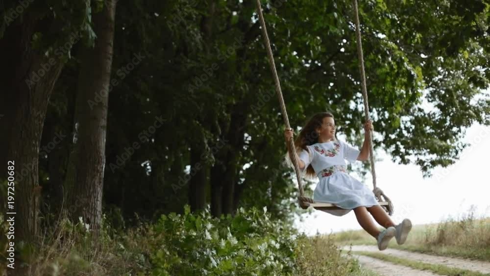 Slow motion of a charming little girl in a white embroidered dress swaying on a wooden swing seat in a garden. Joy of life, moments of happiness. Harmony Slow motion of a charming little girl in a white embroidered dress swaying on a wooden swing seat in a garden. Joy of life, moments of happiness. Harmony