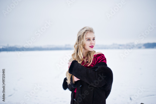 Elegance blonde girl in fur coat and red evening dress posed at winter snowy day.