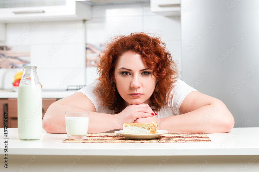 Plus size girl greedy eats sweet pie. Hungry fat model sit by the table and eating dessert. Weight loss concept.  Red curly hair overweight women with delicious cake on kitchen