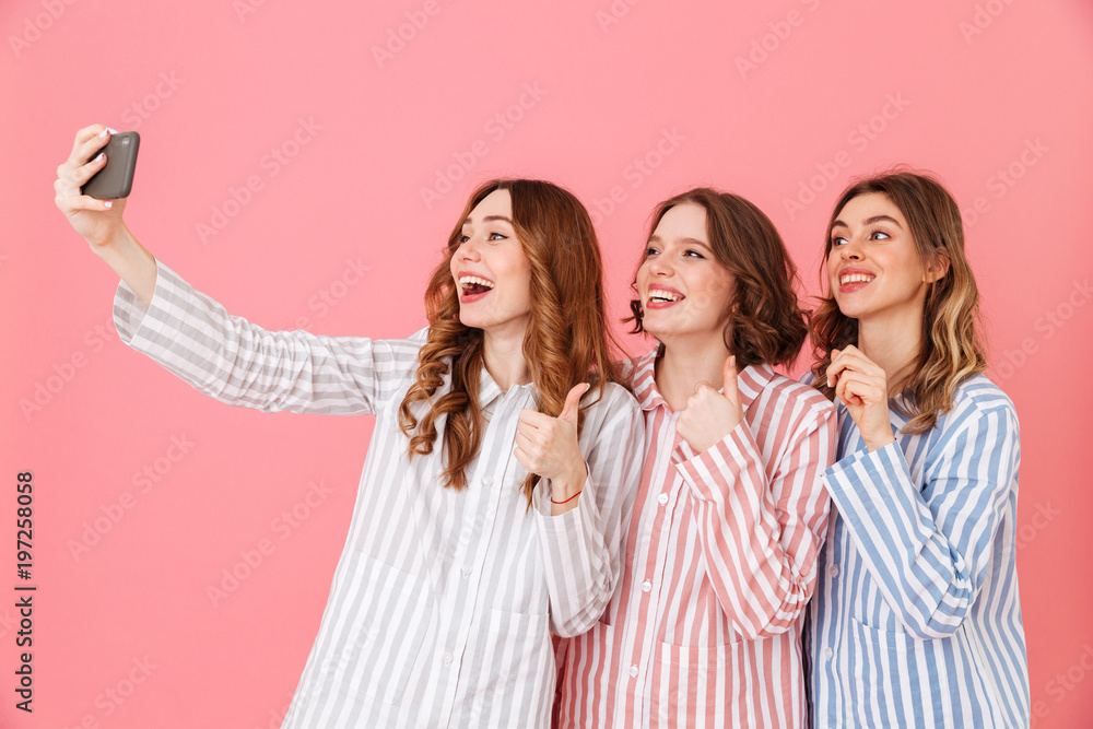 Picture of three cute women 20s wearing colorful striped pyjamas having fun and taking selfie using mobile phone during happy sleepover, isolated over pink background Picture of three cute women 20s wearing colorful striped pyjamas having fun and taking selfie using mobile phone during happy sleepover, isolated over pink background