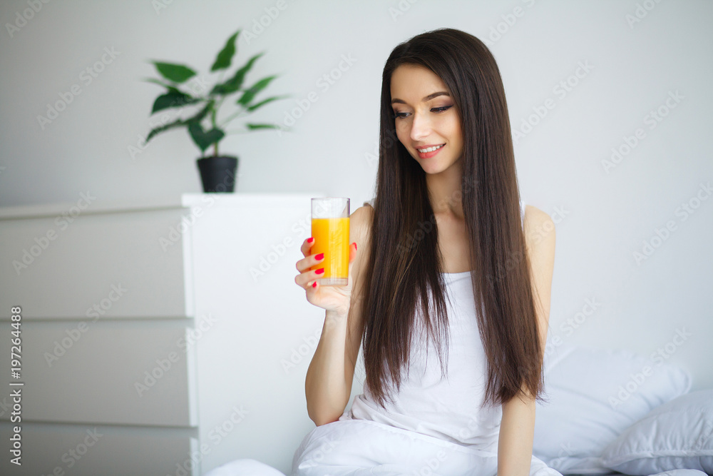 Young woman enjoying a glass of orange juice in the morning as she relaxes sitting on her bed in a healthy diet concept Young woman enjoying a glass of orange juice in the morning as she relaxes sitting on her bed in a healthy diet concept