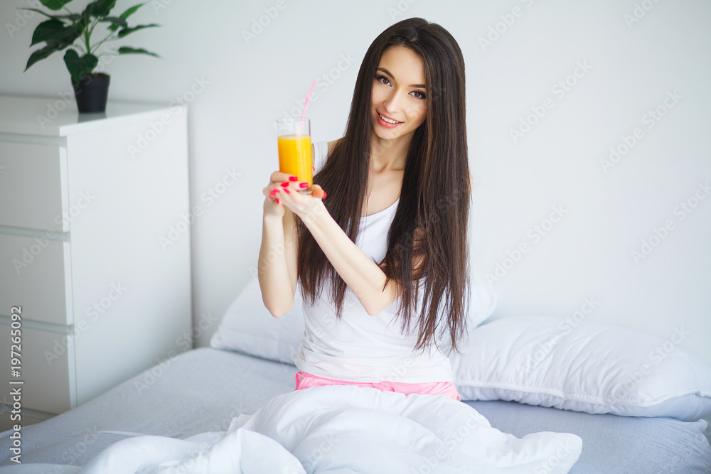 Young woman enjoying a glass of orange juice in the morning as she relaxes sitting on her bed in a healthy diet concept Young woman enjoying a glass of orange juice in the morning as she relaxes sitting on her bed in a healthy diet concept