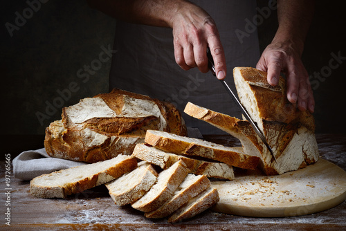 Hands cutting into slices a traditional loaf of freshly baked bread on wooden table in dark setting with grazing light. 