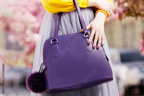 Close up photo of trendy violet bag with fur trinket in hands of fashionable woman posing in street with blooming  spring trees. Fashion elements