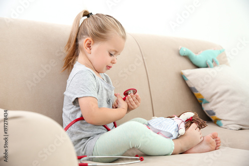 Cute little girl with stethoscope and toy playing on sofa at home