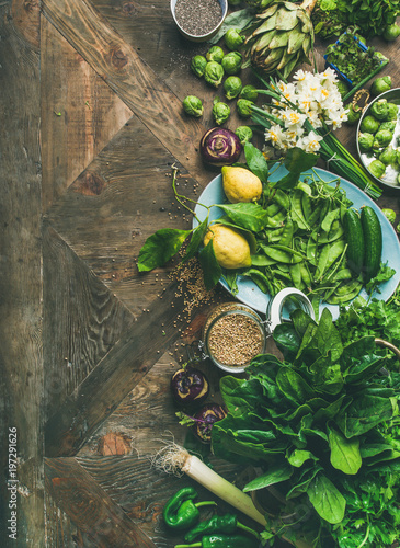 Spring healthy vegan food cooking ingredients. Flat-lay of vegetables, fruit, seeds, sprouts, flowers, greens over wooden background, top view, copy space, vertical composition. Diet food concept