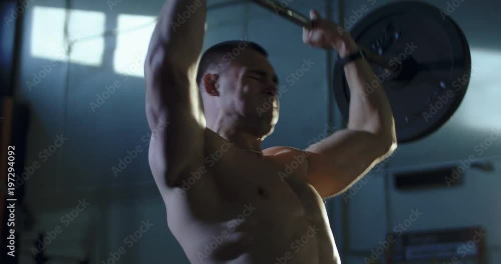 Concentrated athlete without shirt training in gym lifting heavy barbell above head training shoulders. Concentrated athlete without shirt training in gym lifting heavy barbell above head training shoulders.