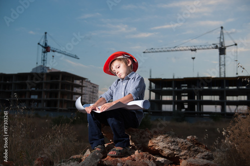 tired little boy wearing helmet solves problem of project on construction site with building crane in the background at end of day