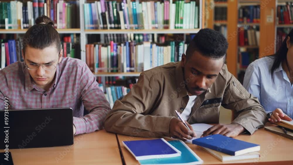 Pan shot of Multi ethnic group of concentrated students preparing for examination while sitting at table in university library Pan shot of Multi ethnic group of concentrated students preparing for examination while sitting at table in university library