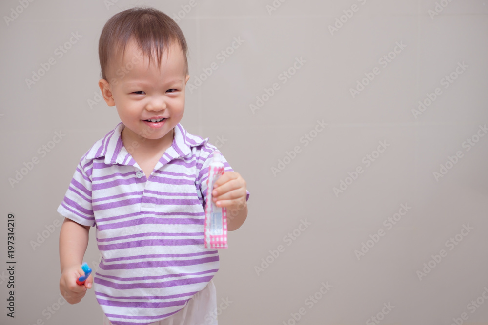 Cute smiling little Asian 18 months / 1 year old toddler boy child wearing purple shirt holding toothbrush and learn to brushing teeth in bathroom at home, Tooth care for children concept
