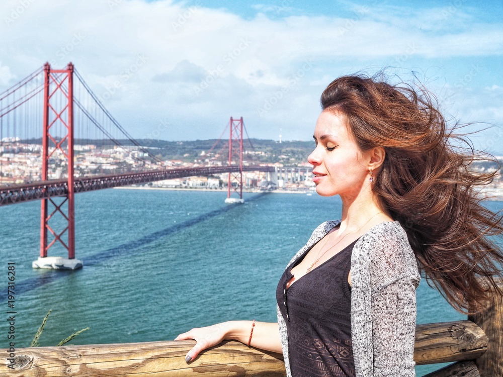 Brunette girl with fluttering in the wind hair stands in the background of the 25th of April bridge in Lisbon. The bridge resembles the Golden bridge in San Francisco