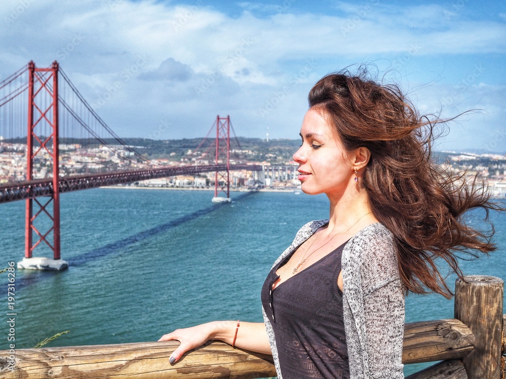 Brunette girl with fluttering in the wind hair stands in the background of the 25th of April bridge in Lisbon. The bridge resembles the Golden bridge in San Francisco Brunette girl with fluttering in the wind hair stands in the background of the 25th of April bridge in Lisbon. The bridge resembles the Golden bridge in San Francisco