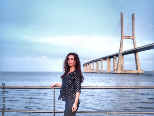 A girl with long hair and in a polka dot shirt stands against the background of the Vasco da Gama Bridge